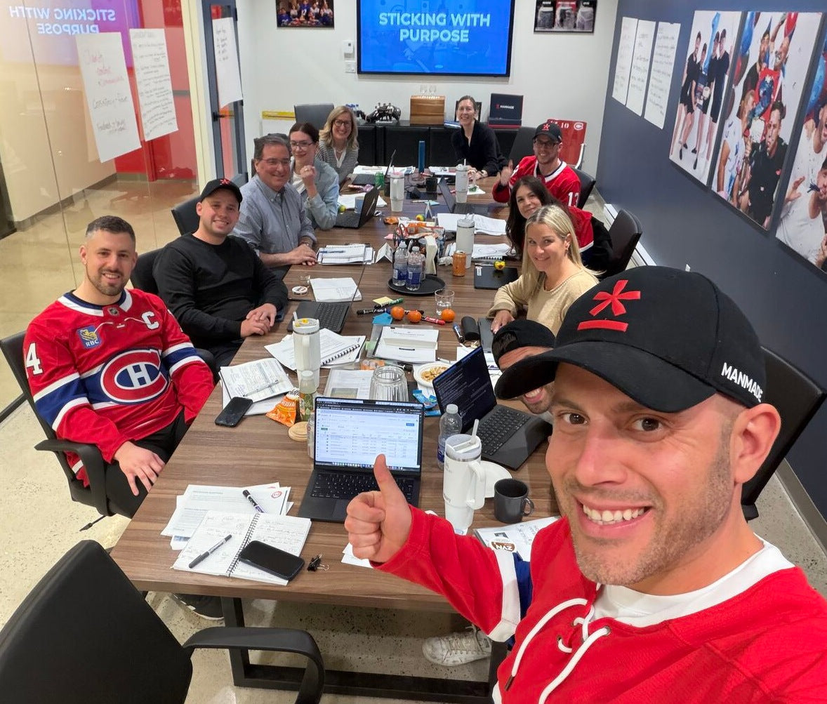 The Manmade team sitting around a conference table in an office, wearing red and white Montreal Canadiens jerseys. In the foreground, Anthony is wearing a black cap with the Manmade logo gives a thumbs-up.