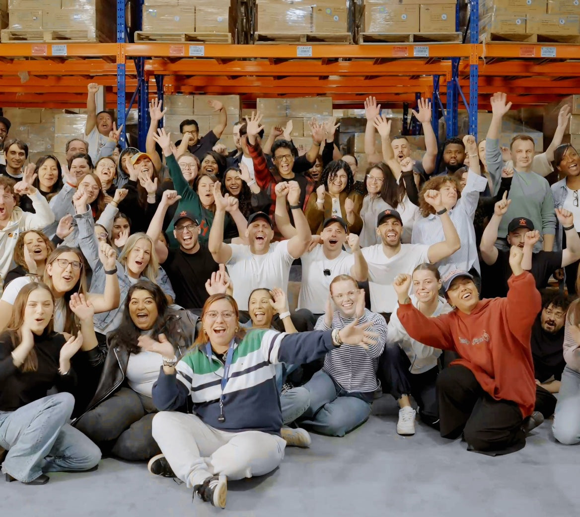 Manmade employees smiling and cheering together in a warehouse filled with cardboard boxes and blue shelving.