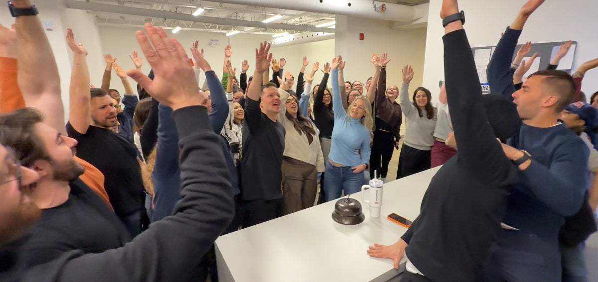 Manmade employees standing indoors with their arms raised in the air, gathered around a white table in a bright office.