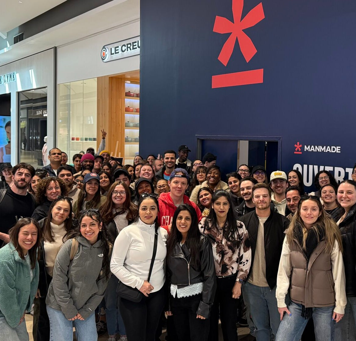 Manmade employees posing for a group photo in a shopping mall in front of the first Manmade retail store, with a prominent dark blue wall behind them featuring our red asterisk logo.