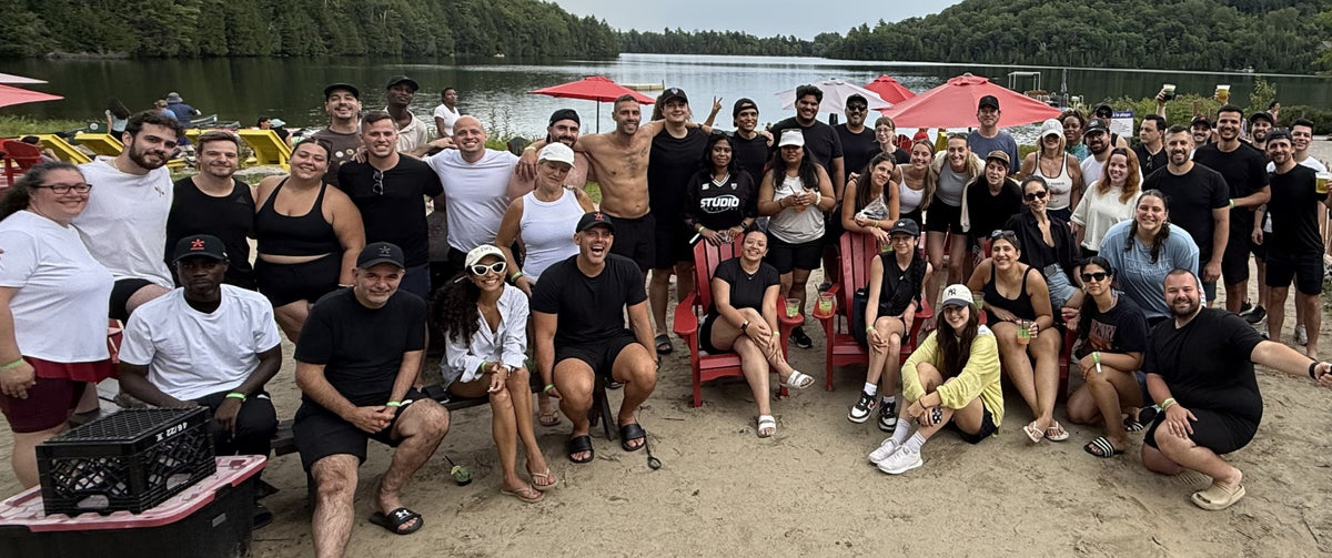 The Manmade team posing together on a sandy beach with water, trees, and red umbrellas in the background.