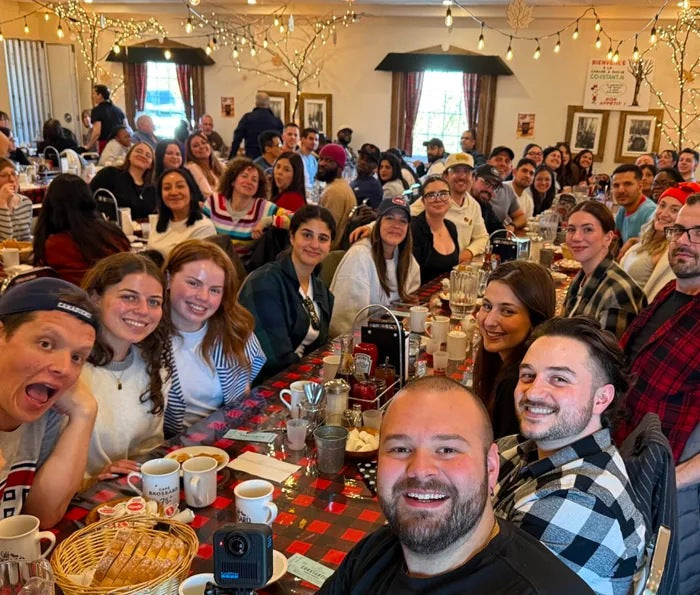 The Manmade team gathered around a long table decorated with a red and black checkered tablecloth, mugs, glasses, and a small camera in the foreground, set in a decorated cabane à sucre with holiday lights.